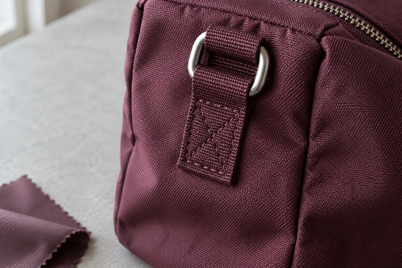 Close-up of a stitched burgundy webbing attachment and metal D-ring on woven bag fabric