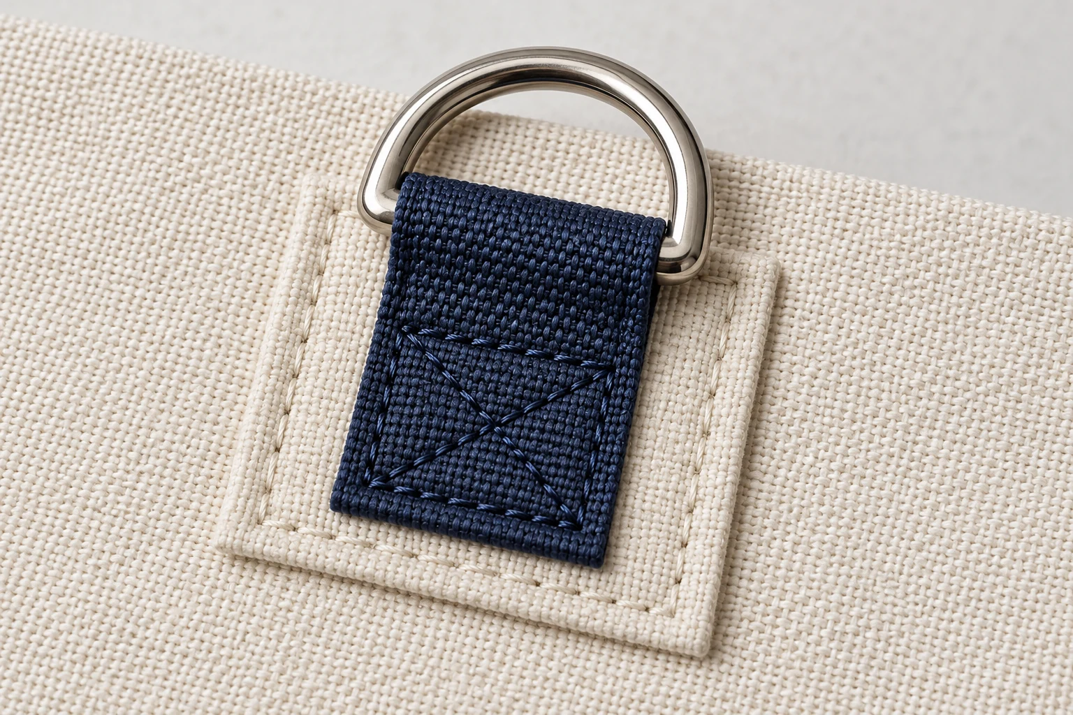 Silver D-ring held by a folded navy webbing tab sewn onto a cream fabric reinforcement patch